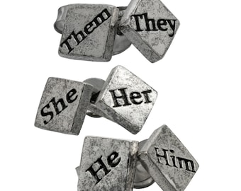 May include: Set of four silver-toned metal earrings with diamond-shaped charms. Each charm has a different pronoun written on it: "She", "He", "They", and "Them".