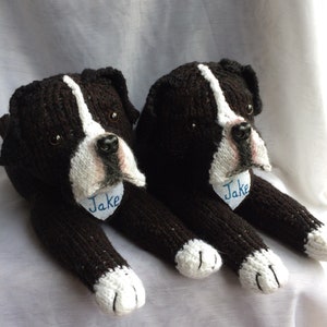 May include: Two black and white knitted dog toys with blue embroidered names "Jake" on their chests. The toys are lying on a white surface.