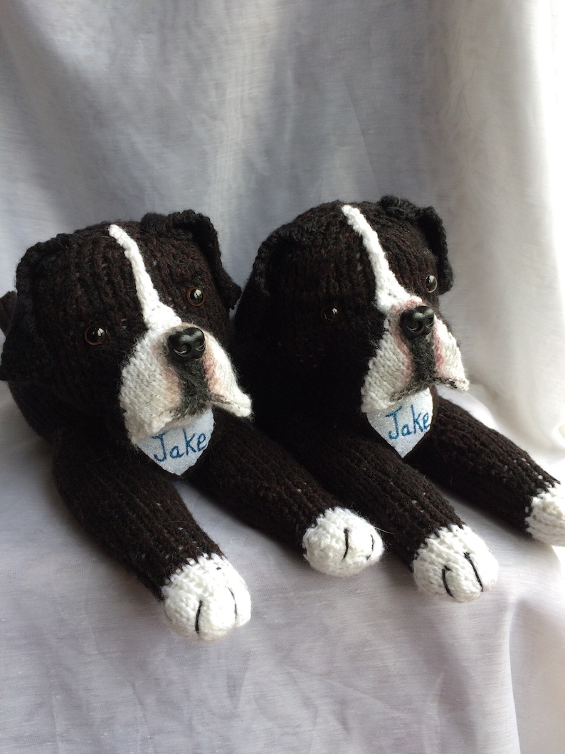 May include: Two black and white knitted dog toys with blue embroidered names "Jake" on their chests. The toys are lying on a white surface.
