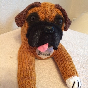 May include: A brown and black knitted dog toy shaped like a boxer dog. The toy has a pink tongue sticking out and a white heart-shaped tag that says "Diesel".