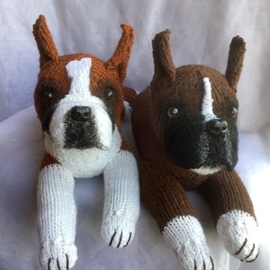 May include: Two brown and white knitted boxer dog plushies. The plushies are lying down with their paws outstretched.