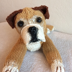 May include: A brown and white knitted dog toy with a brown nose and black eyes. The toy is lying on a white fluffy surface.