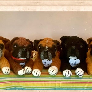 May include: Seven knitted boxer dog plushies, each with a different name tag on a heart-shaped tag. The plushies are arranged in a row on a striped fabric. The names on the tags are:  "Soli", "Joe", "Laika", "Neo", "Millie".