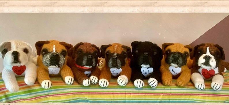 May include: Seven knitted boxer dog plushies, each with a different name tag on a heart-shaped tag. The plushies are arranged in a row on a striped fabric. The names on the tags are:  "Soli", "Joe", "Laika", "Neo", "Millie".