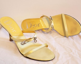 May include: A pair of pale yellow Dior sandals. The sandals feature thin straps, a small heel, and a decorative pin with rhinestones. The Dior logo is visible on the insole. The shoes are on a white surface.