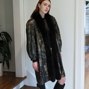 May include: A gold and black patterned coat with a black fur collar. The coat is being worn by a person with red hair and is standing on wooden stairs.