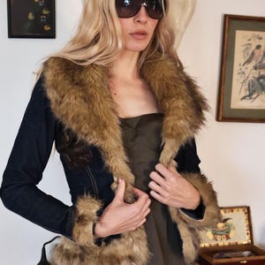 May include: A dark denim jacket with a brown fur collar and cuffs. The jacket has brown leather accents and is worn over a dark green dress. The person is wearing dark sunglasses.