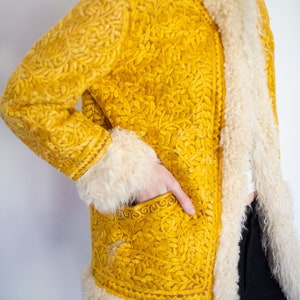 Puede incluir: Una chaqueta amarilla bordada con cuello y ribetes de piel de oveja blanca. La chaqueta tiene un patr&oacute;n floral y un bolsillo en la parte delantera.