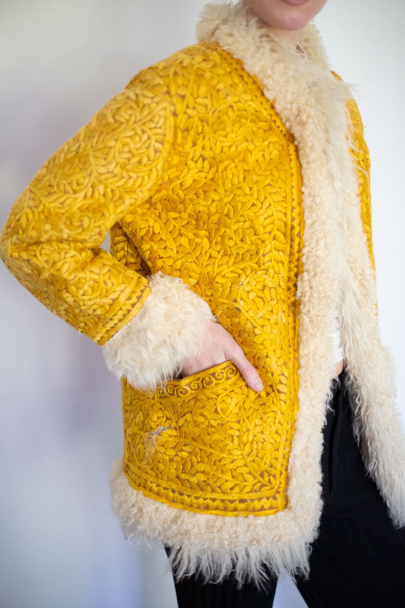 Puede incluir: Una chaqueta amarilla bordada con cuello y ribetes de piel de oveja blanca. La chaqueta tiene un patr&oacute;n floral y un bolsillo en la parte delantera.