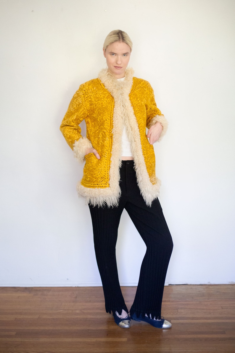 Puede incluir: Una chaqueta de ante amarilla con ribete de piel blanca. La chaqueta tiene una silueta larga y ajustada y un cuello alto.