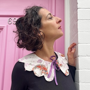 May include: A decorative collar with a white base, featuring a koi fish and floral pattern in shades of red, orange, and purple. The collar has a scalloped edge and a purple ribbon tie. The person is wearing a black top.