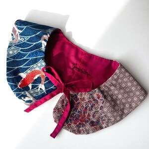 May include: A decorative collar featuring a koi fish design in blue and red, a burgundy interior, and a geometric pattern. The collar has a tie closure and the word "Gracias" embroidered on the burgundy fabric.