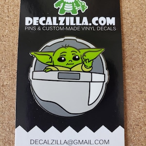 May include: A green and gray enamel pin featuring a cartoon character from the Star Wars franchise, The Mandalorian. The character is sitting in a floating pod with a hand raised in a gesture of greeting. The pin is on a black background with the text "DECALZILLA.COM PINS & CUSTOM-MADE VINYL DECALS" and "DECALZILLA@GMAIL.COM @decalzilla".