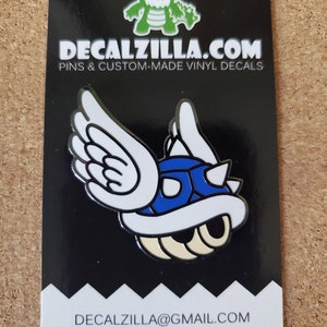 May include: A blue and white enamel pin of a winged Koopa Troopa from the Super Mario Bros. video game series. The pin is on a black background with the text "DECALZILLA.COM PINS & CUSTOM-MADE VINYL DECALS" and "DECALZILLA@GMAIL.COM @decalzilla".