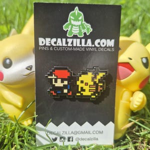 Puede incluir: Una tarjeta negra con un pin pixelado que presenta un personaje y una criatura amarilla. La tarjeta tiene el texto "DECALZILLA.COM" e iconos de redes sociales. Dos figuras amarillas flanquean la tarjeta, sobre hierba verde.