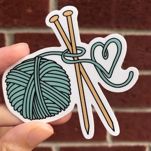 May include: A teal yarn ball with two wooden knitting needles and a heart-shaped loop of yarn.