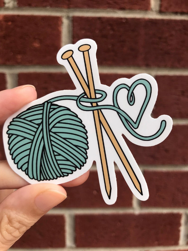 May include: A teal yarn ball with two wooden knitting needles and a heart-shaped loop of yarn.