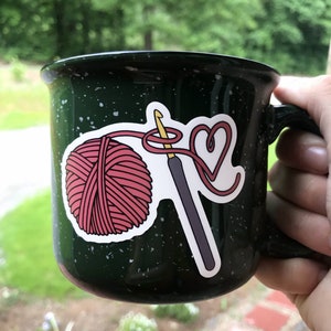 May include: A green camping mug with a white speckled design features a sticker of a pink yarn ball, a crochet hook, and a heart shape made of yarn.