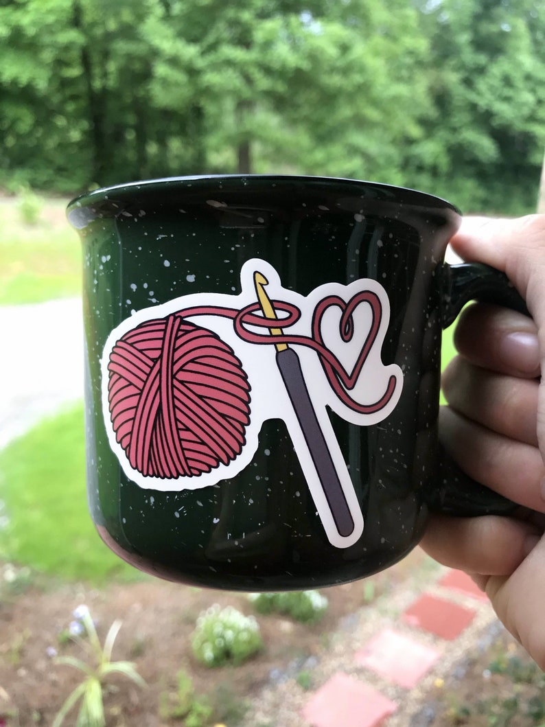 May include: A green camping mug with a white speckled design features a sticker of a pink yarn ball, a crochet hook, and a heart shape made of yarn.