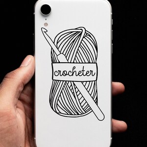 May include: White phone case with a black and white illustration of a ball of yarn and a crochet hook. The yarn ball has a label that says "crocheter".