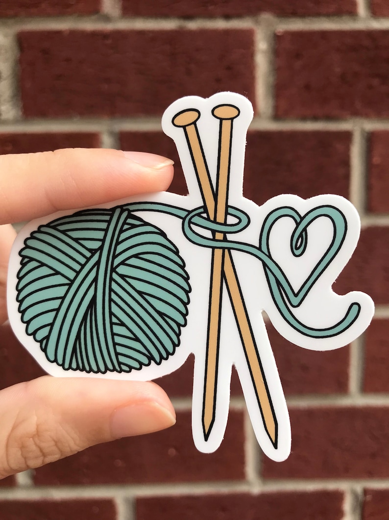 May include: A teal yarn ball with two wooden knitting needles and a heart-shaped loop of yarn.
