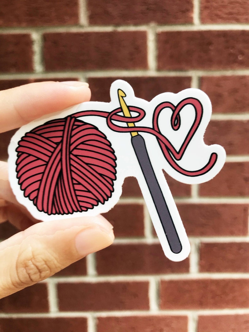 May include: A sticker with a red yarn ball, a gray crochet hook, and a red yarn heart. The yarn is wrapped around the hook and forms the heart shape.
