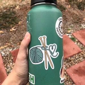 May include: A green reusable water bottle with various stickers, including a yarn ball and knitting needles, a coffee cup with a bicycle wheel, and a floral design.