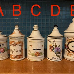 Peut inclure: Cinq pots de pharmacie en céramique blanche avec couvercles, chacun portant l'inscription "Opium". Les pots présentent des motifs floraux et botaniques de différentes couleurs, avec des bordures dorées. Ils sont disposés sur une surface en bois.