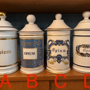 Peut inclure: Quatre pots de pharmacie anciens avec couvercles. Les pots sont blancs avec des bordures dorées et le mot "Opium" en lettres dorées. Deux pots ont des étiquettes décoratives bleues et dorées. Les pots sont exposés sur une surface en bois.