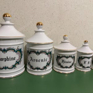 May include: A set of white ceramic apothecary jars with gold detailing. Each jar is labelled with a different substance: Morphine, Arsenic, Heroine, I S, and Opium. The jars are decorated with a floral design and are of varying sizes.