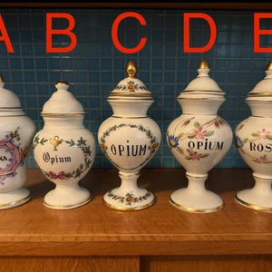 Könnte beinhalten: Fünf weiße Apothekergläser aus Keramik mit goldenen Akzenten und Deckeln. Jedes Glas hat ein anderes Etikett, darunter "COCAINA", "Opium" und "ROSARI:" in schwarzer Schrift. Florale und botanische Designs verzieren die Gläser.