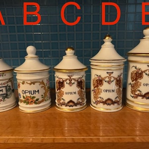 Peut inclure: Cinq pots de pharmacie anciens en céramique blanche avec des garnitures et des couvercles dorés. Chaque pot porte le mot "OPIUM" dans une étiquette rectangulaire. Les pots sont décorés de motifs floraux et botaniques.