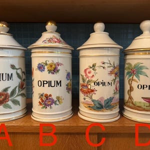 Könnte beinhalten: Vier antike Apothekergläser aus weißer Keramik mit Deckeln, beschriftet mit "OPIUM" in schwarzer Schrift. Jedes Glas hat ein anderes Blumendesign in leuchtenden Farben, mit goldenen Akzenten auf den Deckeln und Rändern. Die Gläser werden auf einem Holzregal ausgestellt.