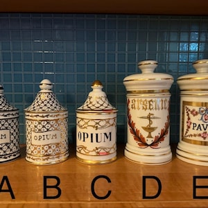May include: Five antique apothecary jars with lids. The jars are white with gold trim and feature labels with the words "OPIUM", "ARSENIC", and "PAVOT". The jars have decorative patterns and are arranged on a wooden surface.