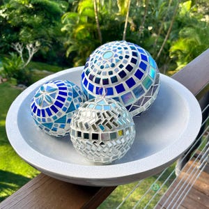 May include: Three mosaic glass spheres in a white bowl. The spheres are various sizes and feature blue, white, and silver mirrored tiles. The bowl is white with a textured surface, and the background shows green foliage.