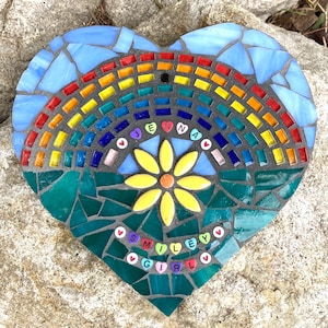 May include: Heart-shaped mosaic art piece with a rainbow design, a yellow flower, and the names "Jenna" and "Smiley Girl" spelled out. The mosaic features blue, green, yellow, orange, and red glass tiles. A hole is at the top for hanging.