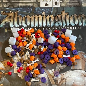 Op de afbeelding: Een verzameling spelstukken voor het bordspel "Abomination: The Heir of Frankenstein". De stukken zijn gemaakt van plastic en zijn verkrijgbaar in verschillende kleuren, waaronder rood, oranje, paars, wit en bruin. De stukken zijn gevormd als botten, schedels, harten en andere anatomische onderdelen.
