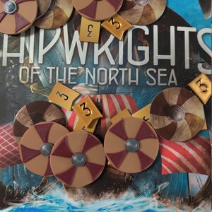 May include: A close-up of a board game with wooden pieces. The pieces are shields with a red cross design and a metal center. The game is called "Shipwrights of the North Sea".