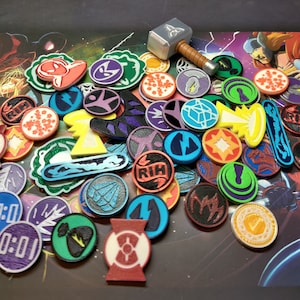 Op de afbeelding: Een verzameling kleurrijke, ronde en stervormige tokens met verschillende ontwerpen, waaronder een hamer, een sneeuwvlok en een bliksem. De tokens zijn gemaakt van plastic en hebben een glanzende afwerking.