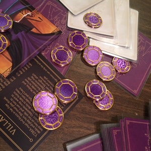 Puede incluir: Fichas de juego moradas y doradas con un dise&ntilde;o geom&eacute;trico. Las fichas est&aacute;n esparcidas sobre una mesa de madera con cartas en el fondo.