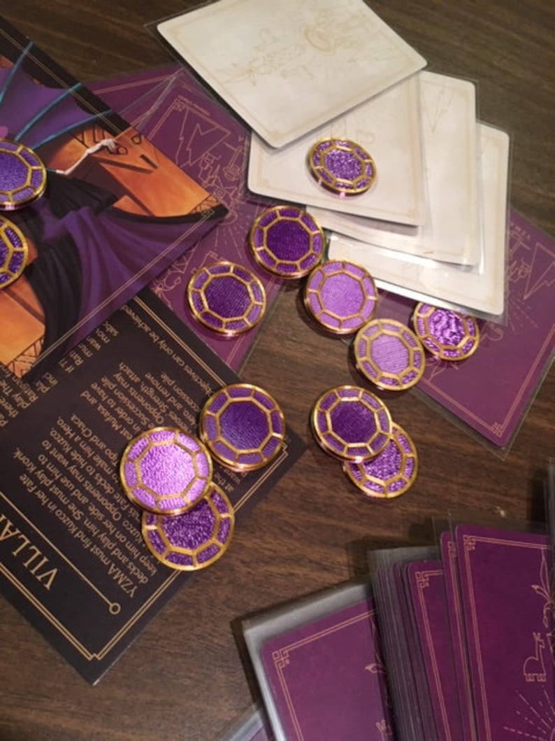 Puede incluir: Fichas de juego moradas y doradas con un dise&ntilde;o geom&eacute;trico. Las fichas est&aacute;n esparcidas sobre una mesa de madera con cartas en el fondo.