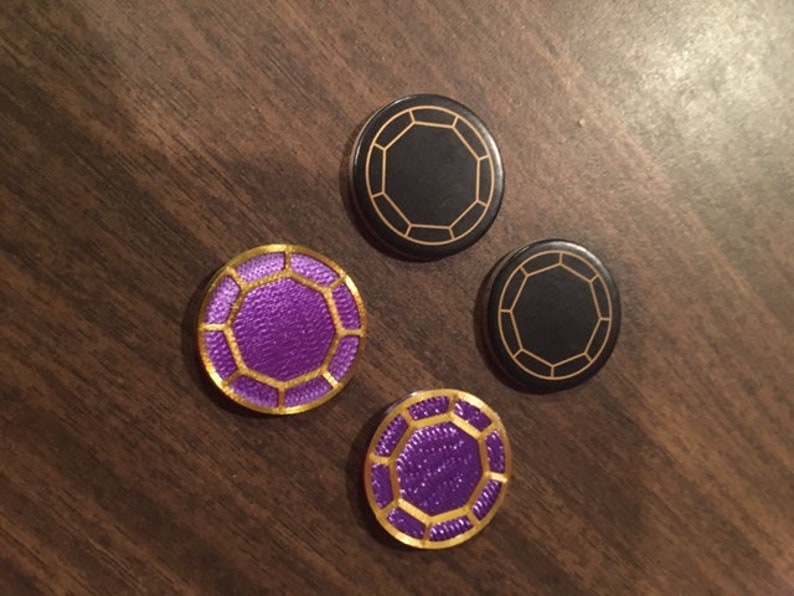 Puede incluir: Cuatro fichas de juego redondas de forma octogonal. Dos fichas son negras con bordes dorados y dos fichas son moradas con bordes dorados.