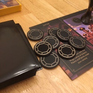 Puede incluir: Primer plano de un tablero de juego con fichas negras y doradas. El tablero presenta el texto "Jafar" y "Magic Lamp at Sultan's Palace and Genie under your control."