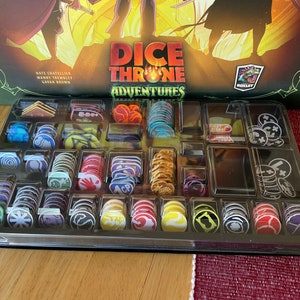 May include: A close-up of a game box organiser filled with colourful game pieces. The organiser is labelled "Dice Throne Adventures".