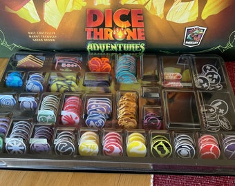 May include: A close-up of a game box organizer filled with colorful game pieces. The organizer is labeled "Dice Throne Adventures".