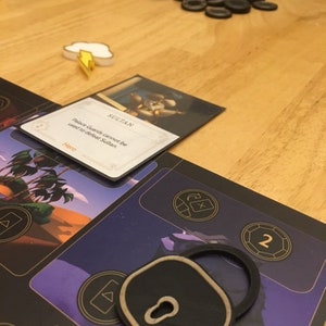 Puede incluir: Un token de cerradura negro con un ojo de cerradura dorado y un borde dorado. El token est&aacute; en un juego de mesa con un fondo morado y el texto "CAVE OF W" en letras doradas. El texto "SULTAN" es visible en una tarjeta en el fondo.