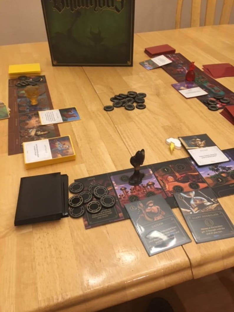 Puede incluir: Un tablero de juego con piezas de juego, cartas y fichas negras y doradas. El tablero de juego presenta un mapa con diferentes ubicaciones y un reloj de arena negro y dorado. El juego se llama "Guildhall".