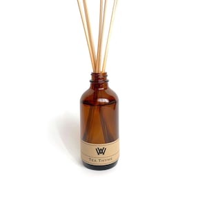 May include: A brown glass bottle with a brown label that says "TEA THYME" and a black "W" logo. The bottle is filled with a clear liquid and has wooden sticks sticking out of the top.