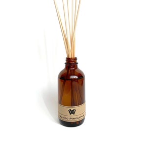 Könnte beinhalten: Eine braune Glasflasche mit einem Etikett mit der Aufschrift "Mango Pineapple" und einem weißen Logo. Die Flasche ist mit einer klaren Flüssigkeit gefüllt und hat Holzstäbchen, die aus der Oberseite herausragen.