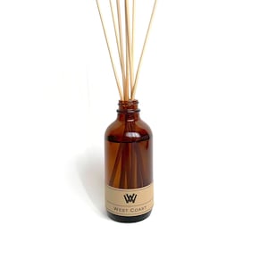 Op de afbeelding: Een bruine glazen fles met een etiket waarop "West Coast" en een gestileerd "W"-logo staan. De fles is gevuld met een heldere vloeistof en heeft houten stokjes die uit de bovenkant steken.